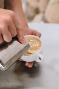Two hands holding a steaming pitcher and a cup with a latte in it, showing latte art